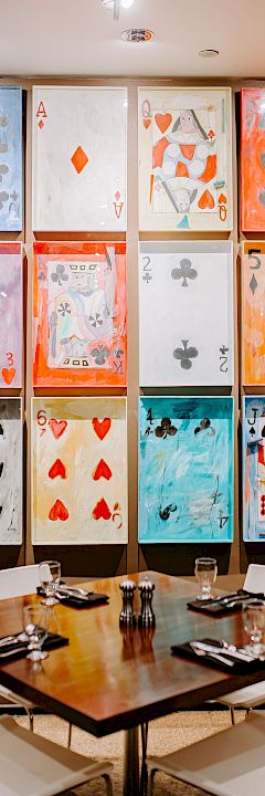 A dining table set for four with utensils and glasses, under a wall display of large, colorful playing card artwork arranged in a 3x4 grid.