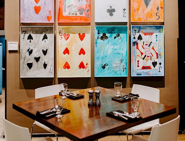 A dining table set for four with utensils and glasses, under a wall display of large, colorful playing card artwork arranged in a 3x4 grid.