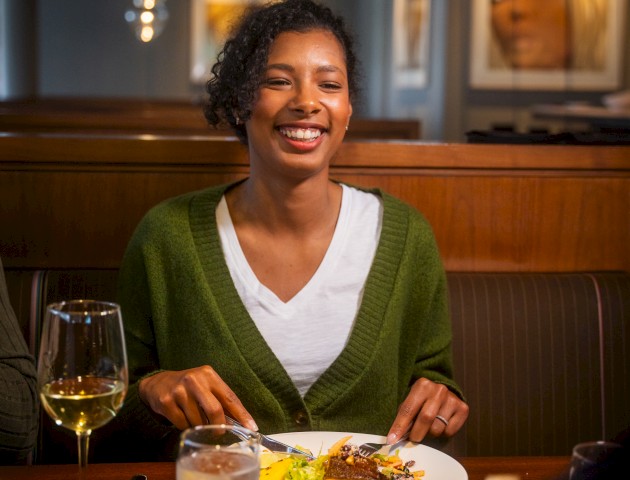 A woman is sitting at a restaurant table, smiling with a plate of food in front of her, and there is a glass of white wine and water on the table.