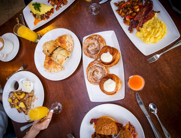 The image showcases a table with assorted breakfast foods, including pastries, eggs, bacon, biscuits, and drinks like orange juice and coffee.