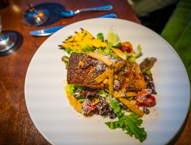 The image shows a plated meal with grilled fish on a bed of mixed greens, cherry tomatoes, and garnished with grilled pineapple slices and lime.