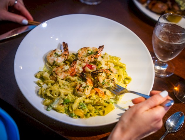 A person is about to eat a shrimp pasta dish with a drink beside them on the table, using a knife and fork.