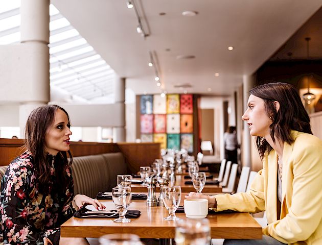 Two women are sitting at a restaurant table, engaged in conversation. The restaurant has modern decor and several glasses are on the table.