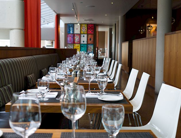 A modern restaurant interior with neatly arranged dining tables set with glassware, plates, and utensils, featuring colorful wall art at the back.