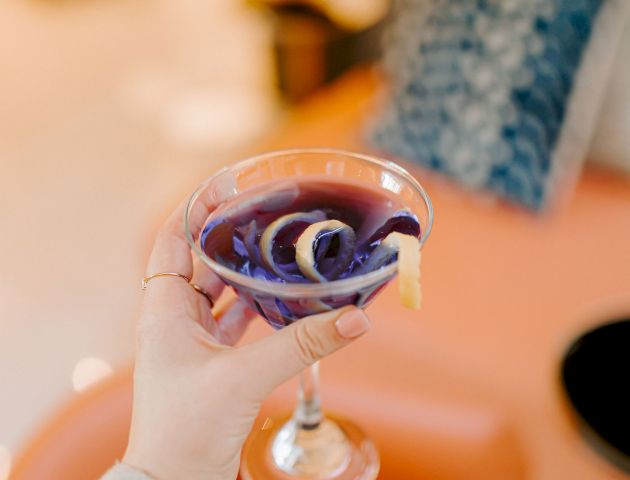 A hand holds a purple cocktail in a glass, garnished with lemon zest curls, with a blurred background of furniture and pillows.