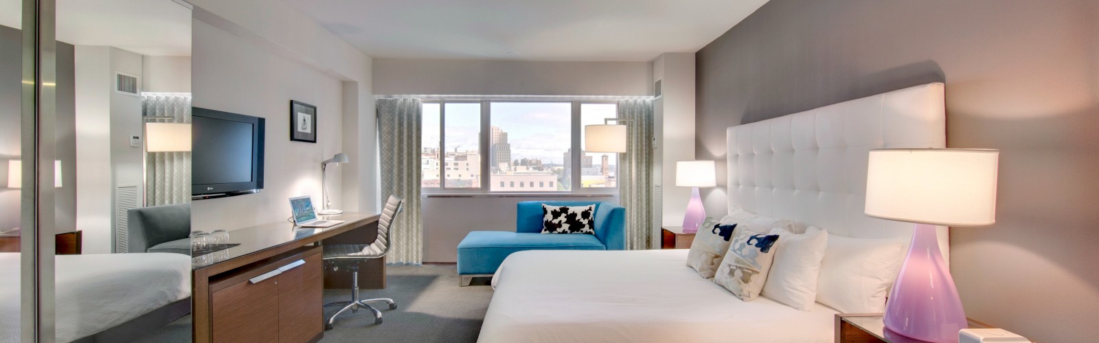 A modern hotel room features a bed, desk, TV, and a window with city views, decorated in neutral tones with purple lamps.