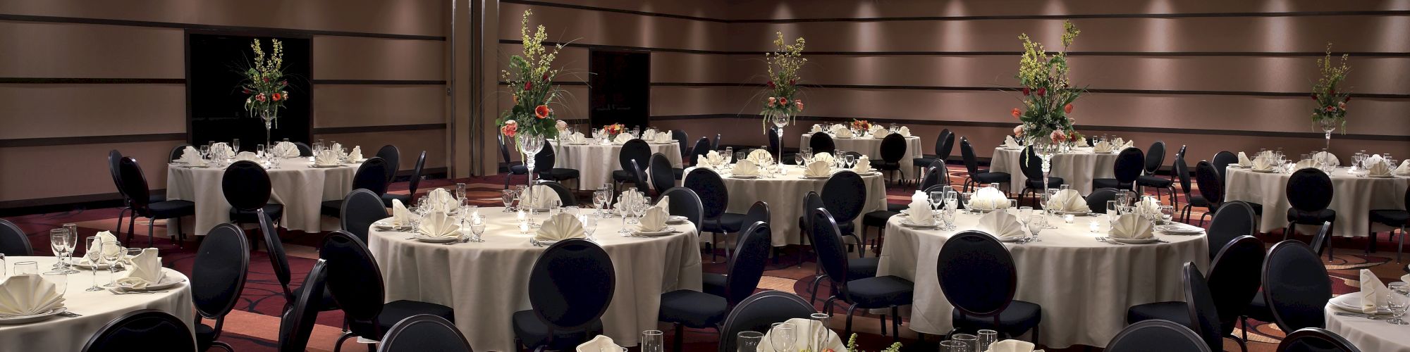 The image shows a large, elegantly decorated banquet hall with round tables set with white tablecloths, napkins, glasses, and floral centerpieces.