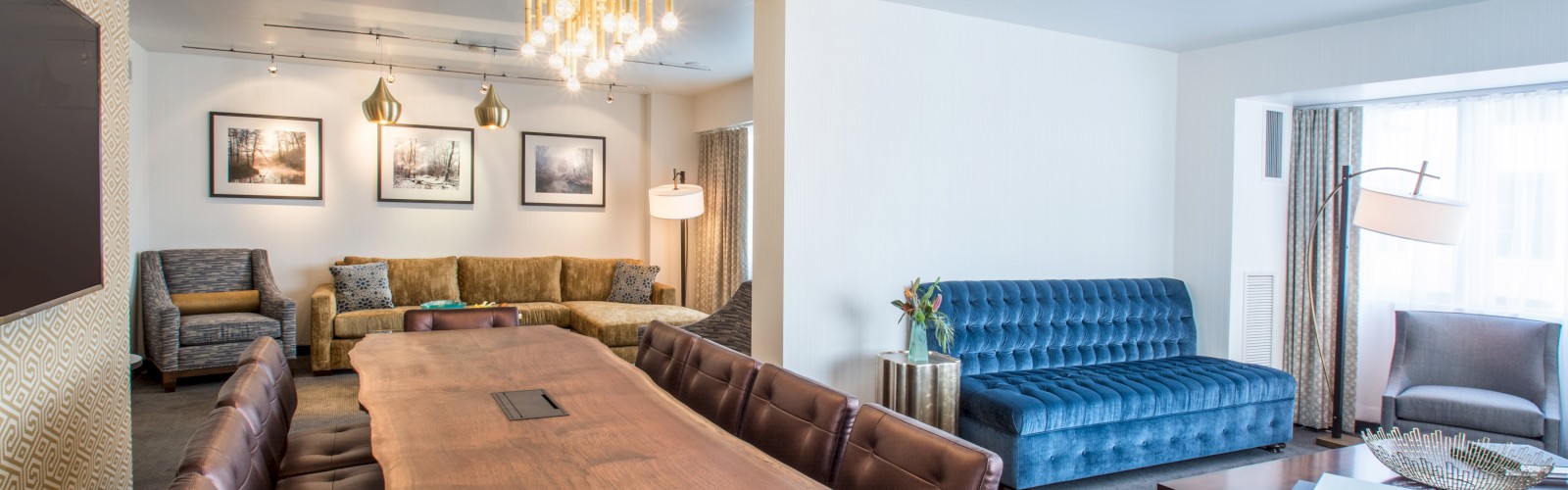 The image shows a modern living room with a long wooden dining table, brown leather chairs, a blue sofa, beige couch, and a chandelier.