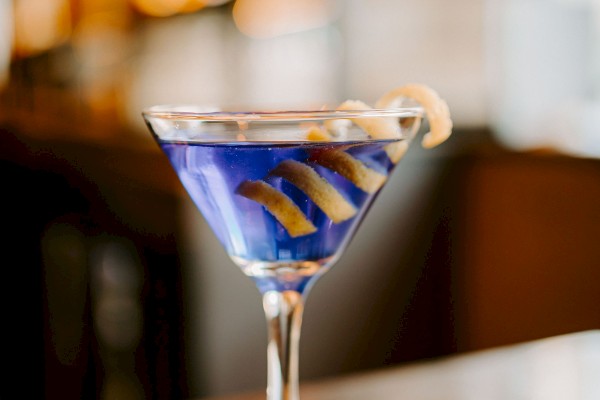 A blue cocktail in a martini glass with a lemon twist sits on a coaster, against a blurred background.