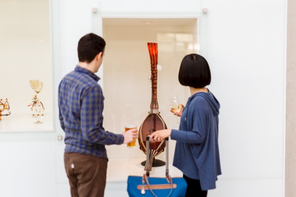 Two people are viewing a tall, decorative vase in a glass display case, each holding a drink in their hands.