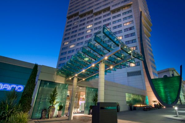 The image shows a modern building with glass entrance canopy, illuminated at dusk, featuring contemporary design elements and greenery.