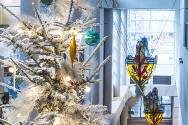 A decorated Christmas tree with lights and ornaments is in a modern indoor setting with colorful hanging decorations.