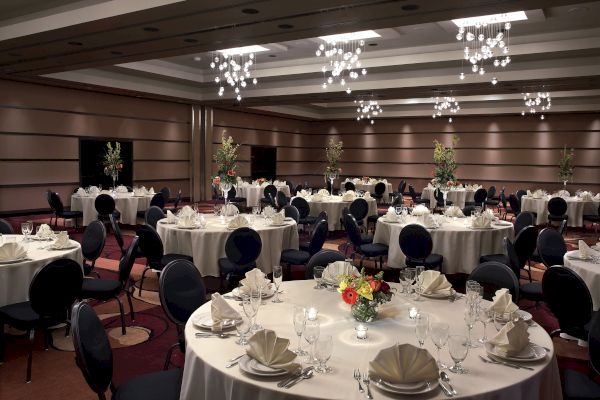 A banquet hall set for an event with round tables, elegant table settings, floral centerpieces, and chandeliers overhead, creating a formal ambiance.