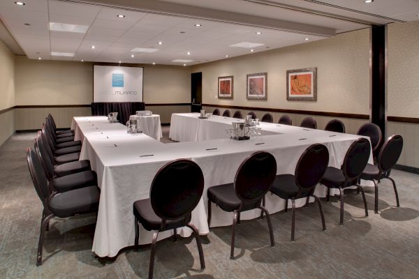 A conference room set up with a U-shaped table arrangement, black chairs, a projector screen, and framed artwork on the walls.