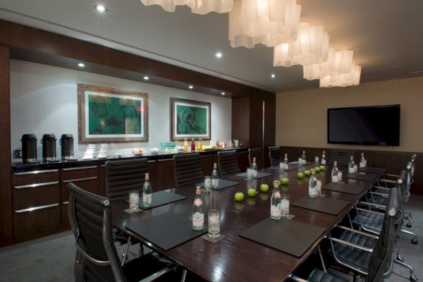 A modern conference room with a long table, black chairs, water bottles, green apples, a TV screen, and artwork on the wall ends the sentence.