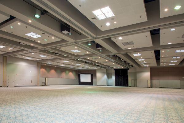 This is an image of an empty, large convention or banquet hall with a carpeted floor, high ceiling, and a projection screen at the far end of the room.