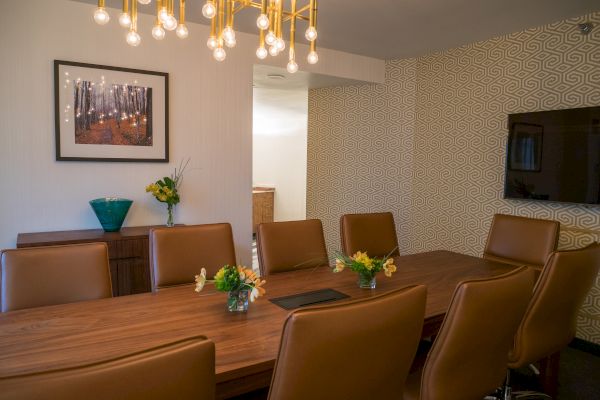 A dining room with a wooden table, brown chairs, a modern light fixture, wall art, vases with flowers, and a wall-mounted TV features.