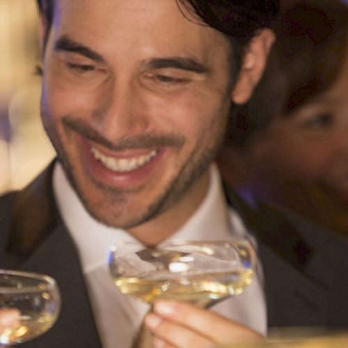 A man and woman smiling while holding champagne glasses in a celebratory toast, with a blurred background and warm lighting.
