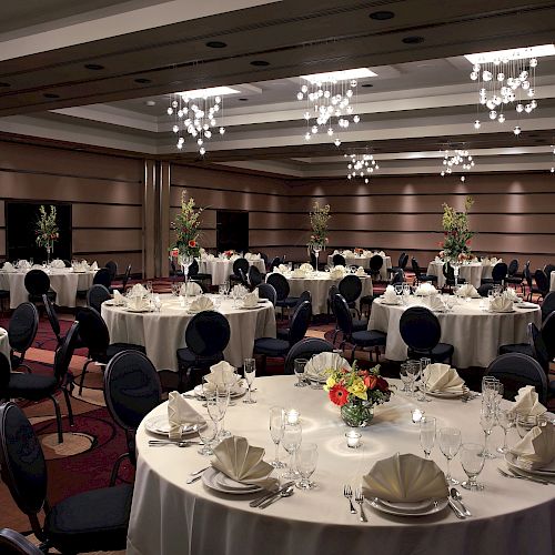 A decorated banquet hall with round tables set for an event, featuring floral centerpieces, folded napkins, glasses, and elegant chandeliers above.