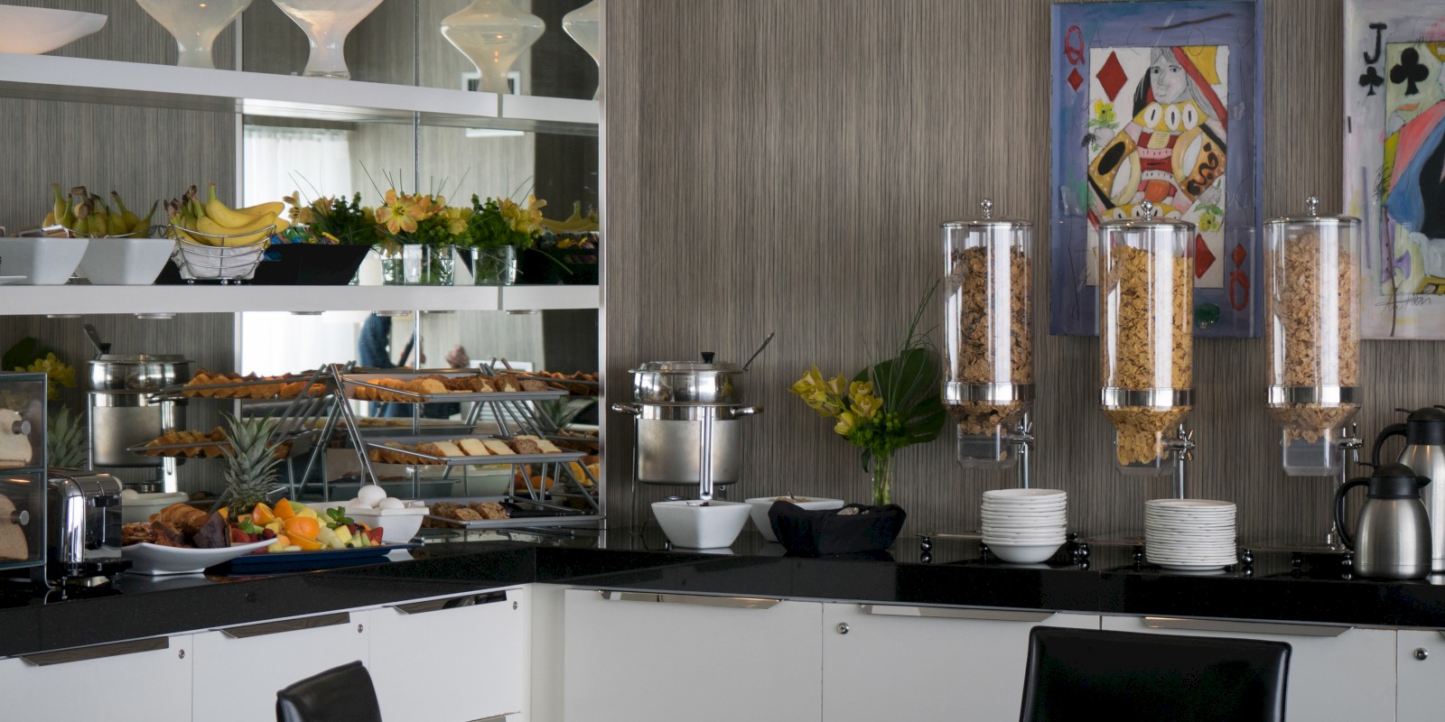 A modern breakfast buffet setup with cereal dispensers, a coffee maker, various fruits, pastries, and decorative items, in a clean dining room.