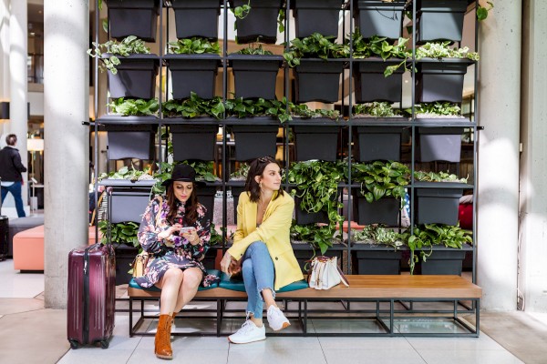 Two people sit on a bench in front of a vertical garden, with one looking at their phone and the other gazing away, luggage beside them.