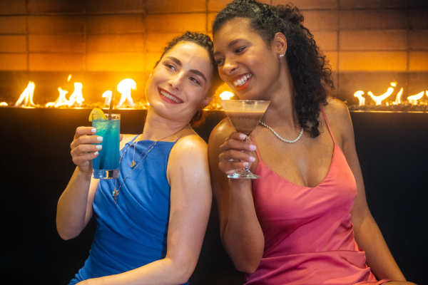 Two women in elegant dresses enjoy cocktails while sitting in front of a fireplace, smiling and posing for the camera.