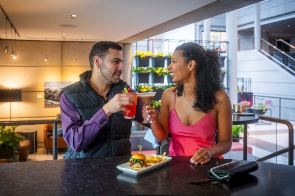 A couple is toasting with drinks at a bar, smiling at each other. A burger and fries are on the table in front of them.