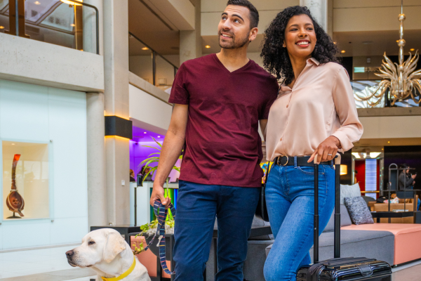 A couple stands in an elegant lobby with a guide dog and a suitcase, looking happy and ready for their journey.