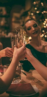 A group of four women is toasting with champagne glasses in a festive, warmly lit setting, possibly celebrating at a restaurant or bar during the holidays.