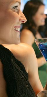 A woman holding a blue cocktail and smiling with other women in the background at what appears to be a social gathering or event.