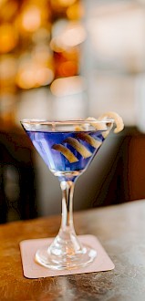 A cocktail in a martini glass with a blue liquid and lemon twist garnish sits on a coaster on a bar counter.