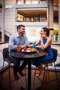 A man and woman are sitting at a table in a modern setting, enjoying drinks and a meal, and engaging in conversation.