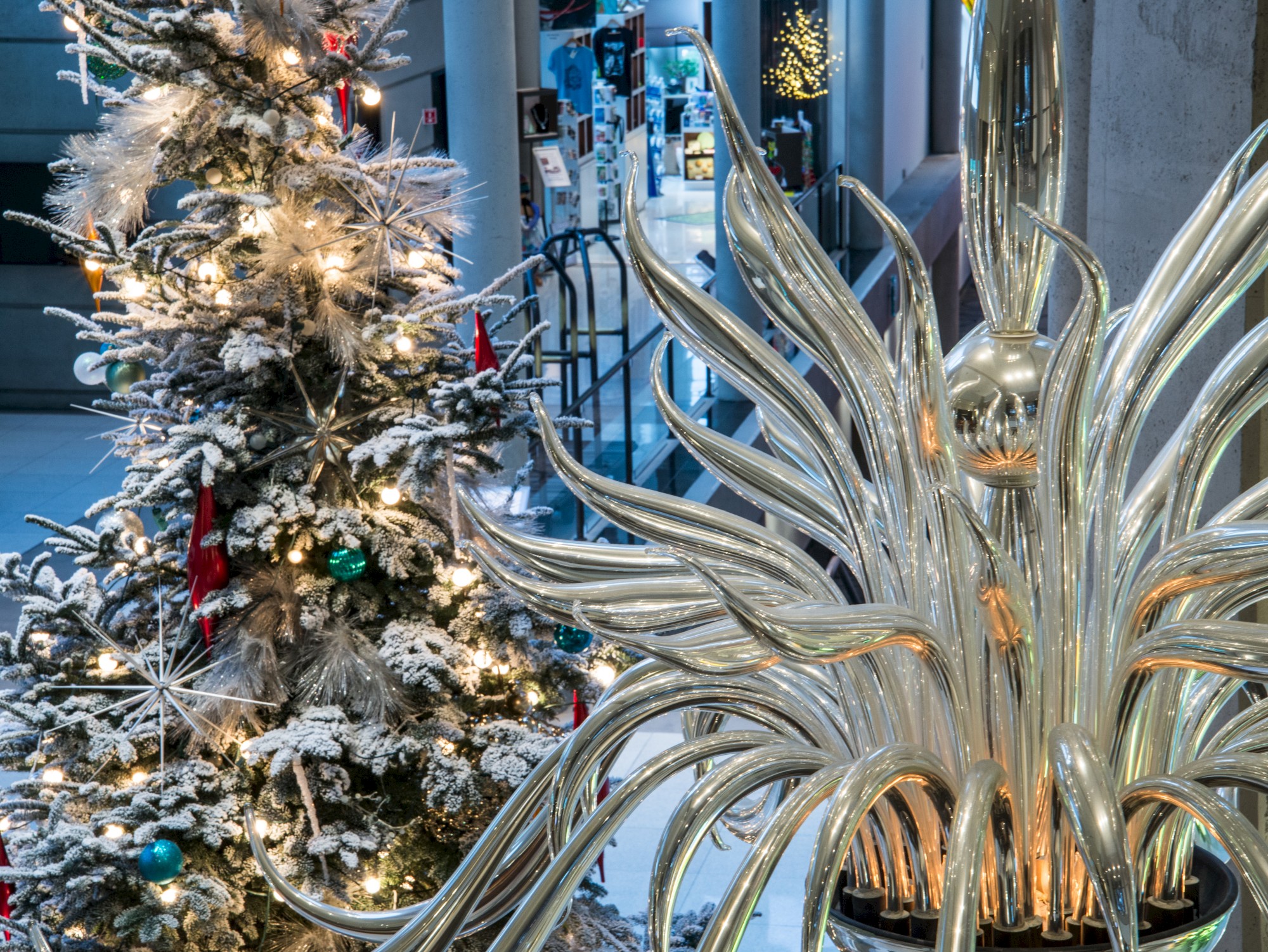 A Christmas tree with ornaments and lights next to a shiny, abstract metallic sculpture inside a modern building.