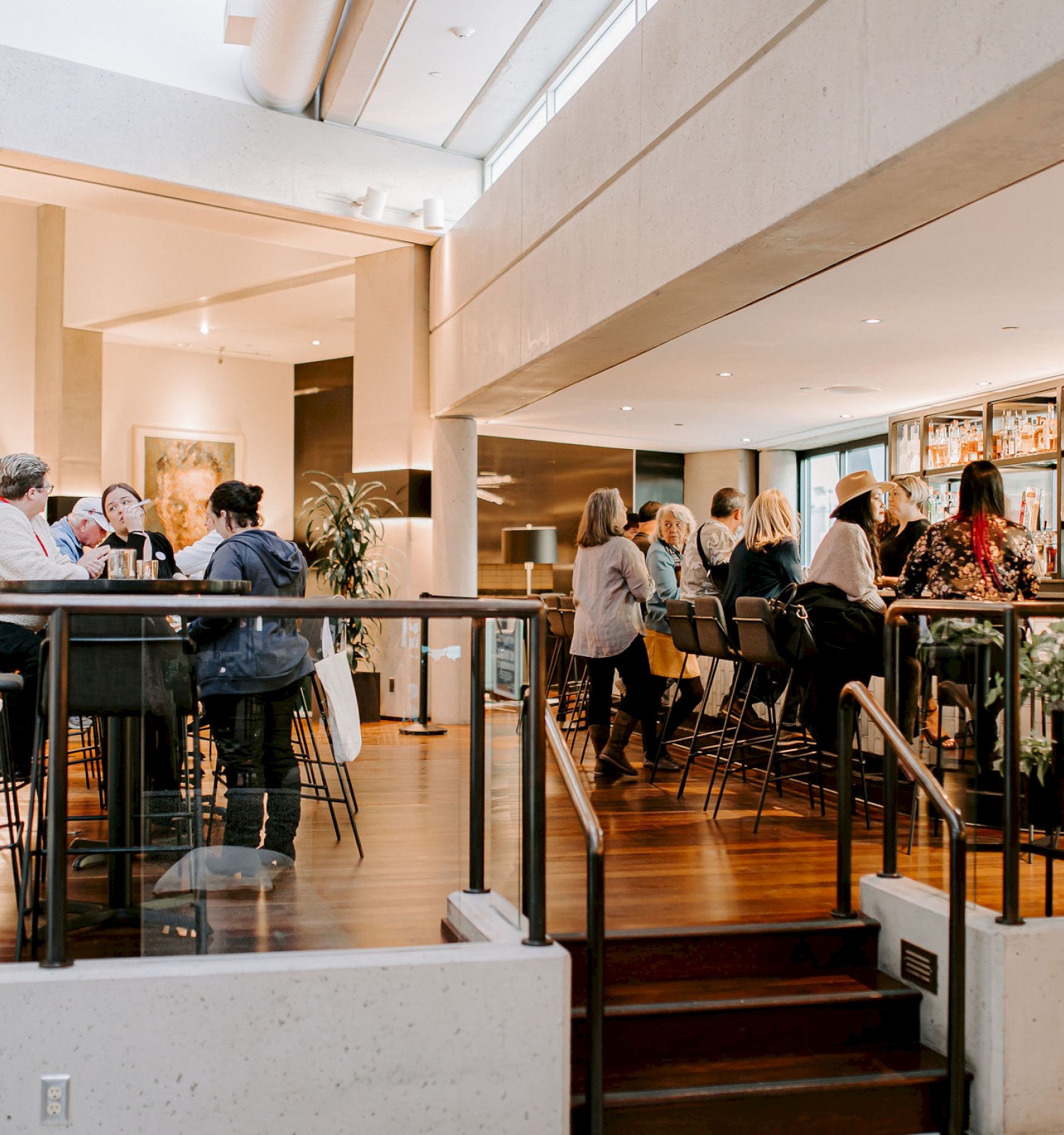 People socializing in a modern, well-lit café or bar with high ceilings and wooden floors. A child walks by in the foreground.