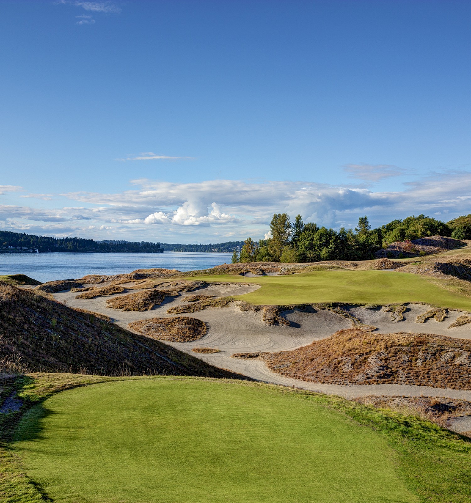 A scenic golf course with rolling hills and sand traps, bordered by water and green trees under a clear blue sky.