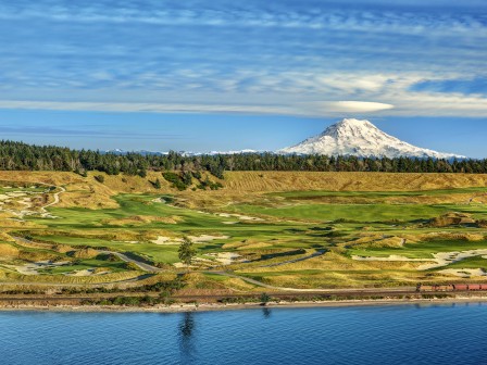 A golf course by a body of water, with a snow-capped mountain in the background under a partly cloudy sky.