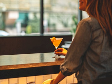 A person seated at a bar, holding a cocktail, with a blurred view outside through large windows in the background.