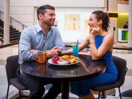 A couple enjoys drinks and snacks at a modern indoor café, sitting across from each other and smiling warmly.