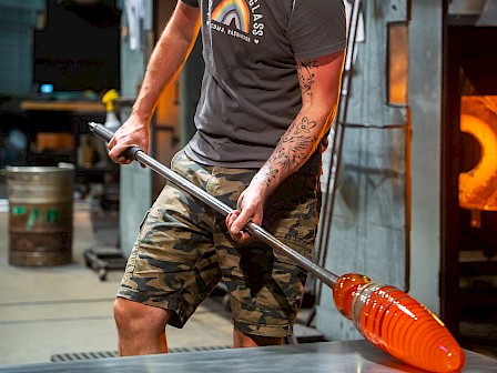 A person works with molten glass on a metal rod in a glassblowing studio, wearing a gray shirt and camouflage shorts.