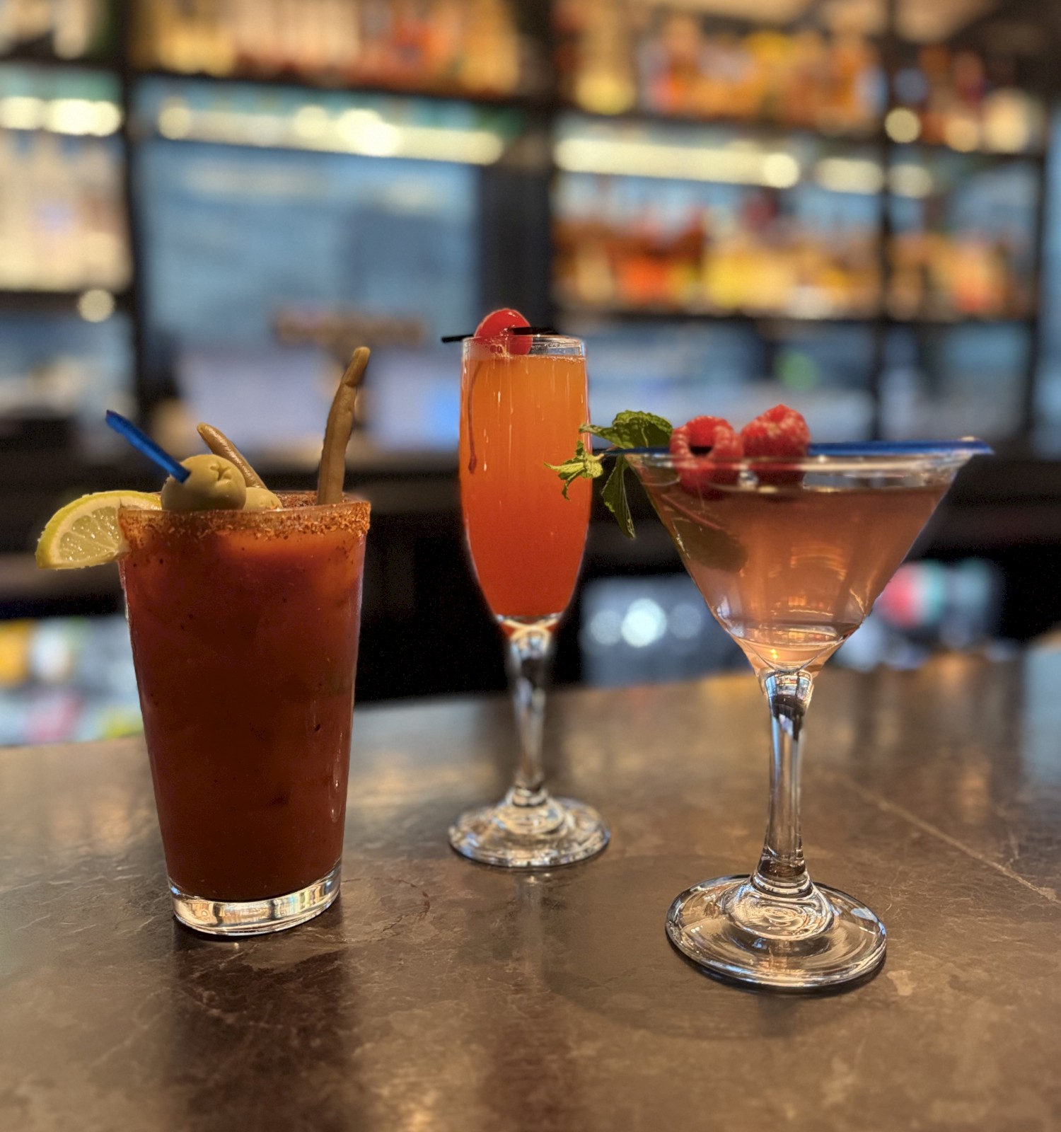 Three cocktails on a bar counter, garnished with fruit and herbs, and a blurry backdrop of shelves and lights completes the scene.