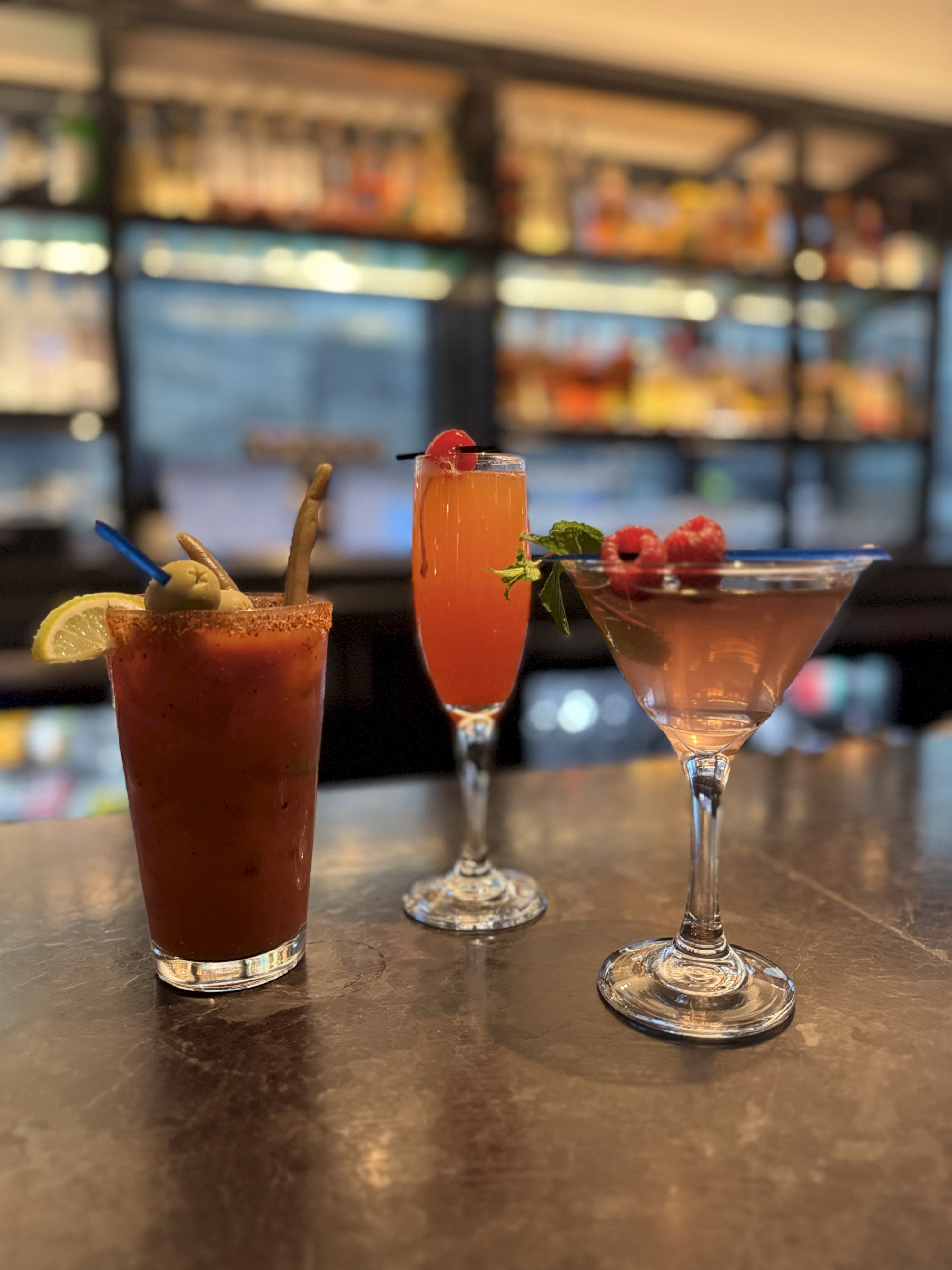 Three cocktails on a bar counter, each garnished differently, with a blurred background of shelves.