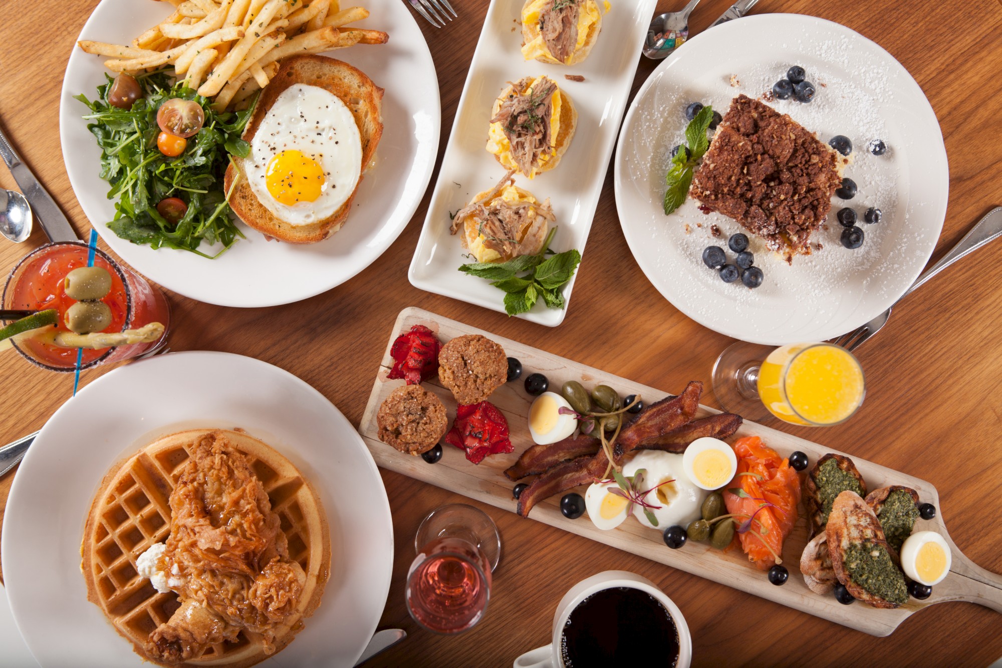 A brunch spread with fried chicken waffles, eggs, toast, salad, fruit, assorted pastries, drinks, and more on a wooden table.