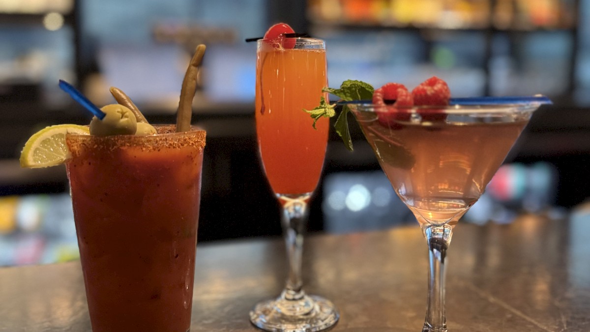 Three cocktails on a bar counter, garnished with fruit and herbs, and a blurry backdrop of shelves and lights completes the scene.