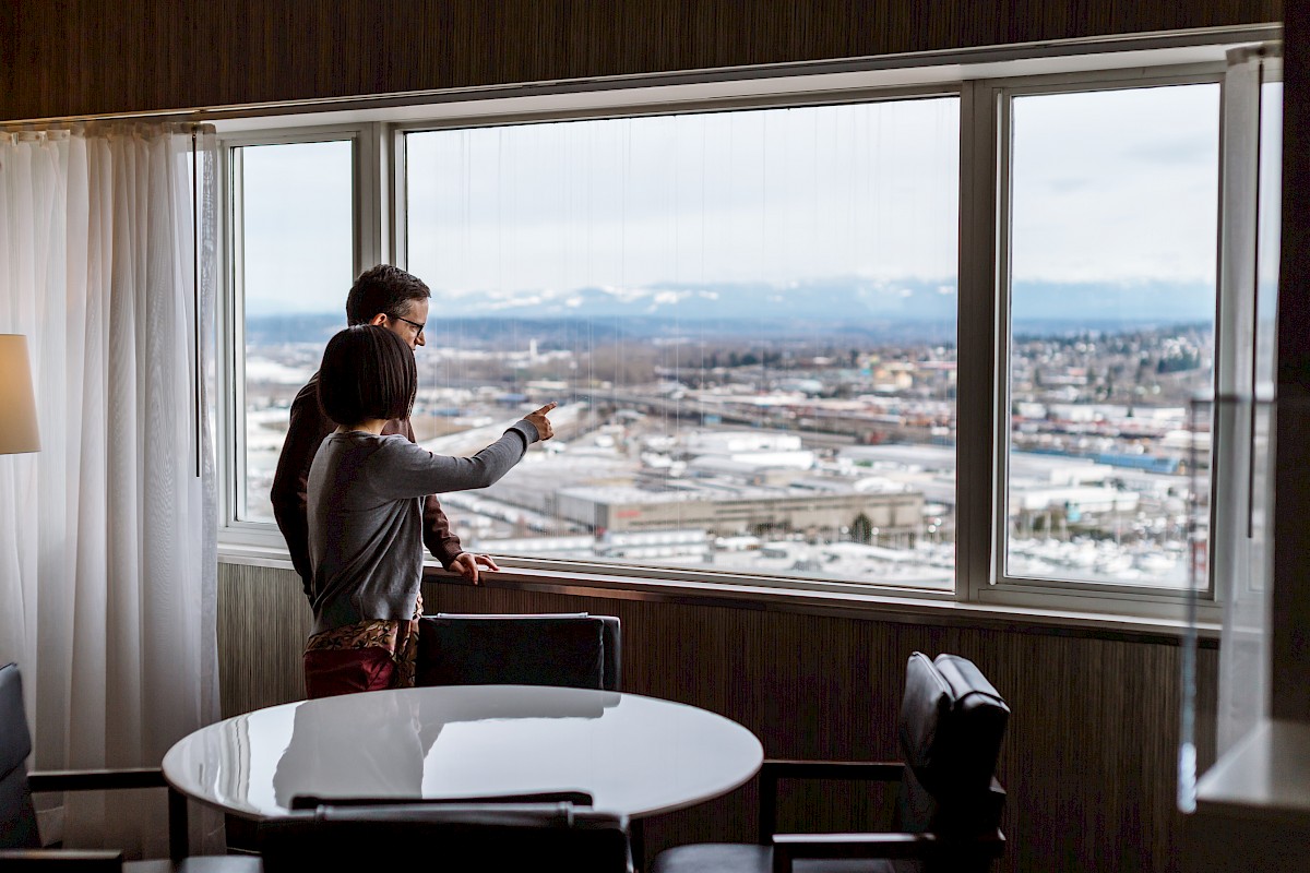 Two people stand by a large window, overlooking an urban landscape with mountains in the distance, inside a room with a table and chairs.