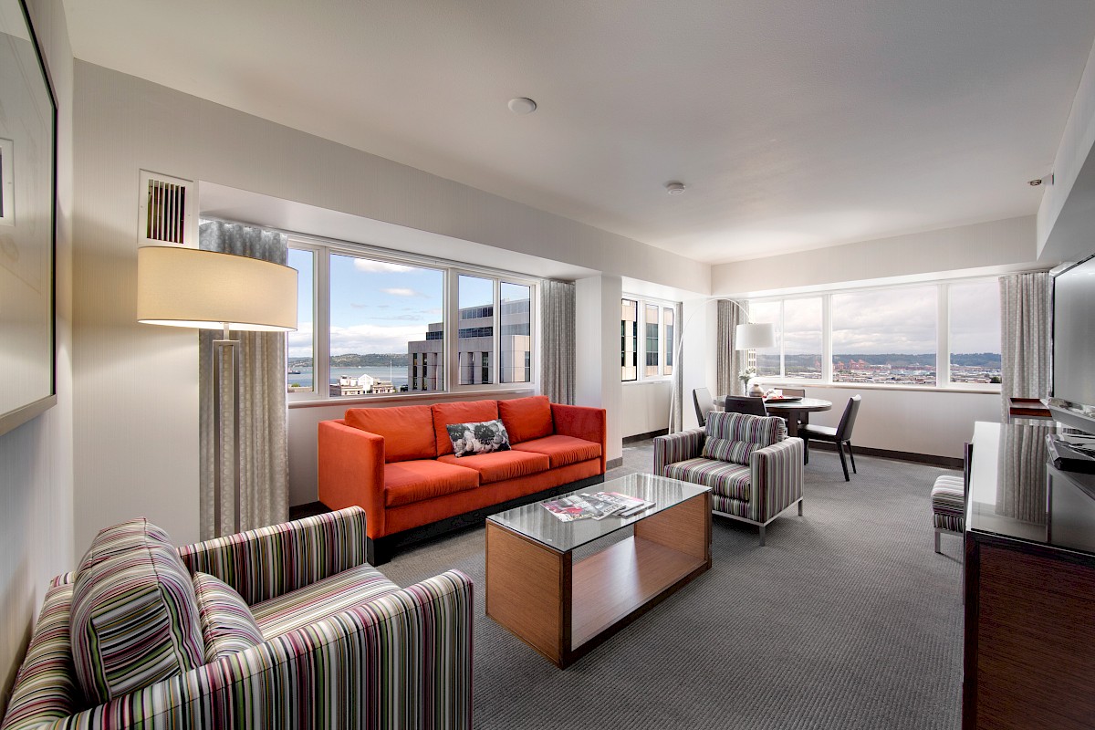 The image shows a living room with a red sofa, striped chairs, a glass coffee table, large windows, and a city view.