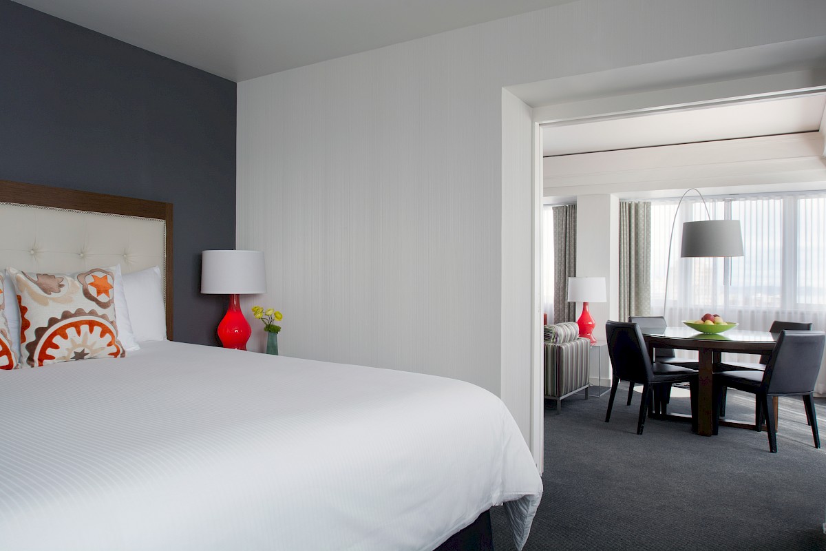 A modern hotel room features a bed with colorful pillows, a lamp, and a seating area with a table and chairs visible in the adjoining room.