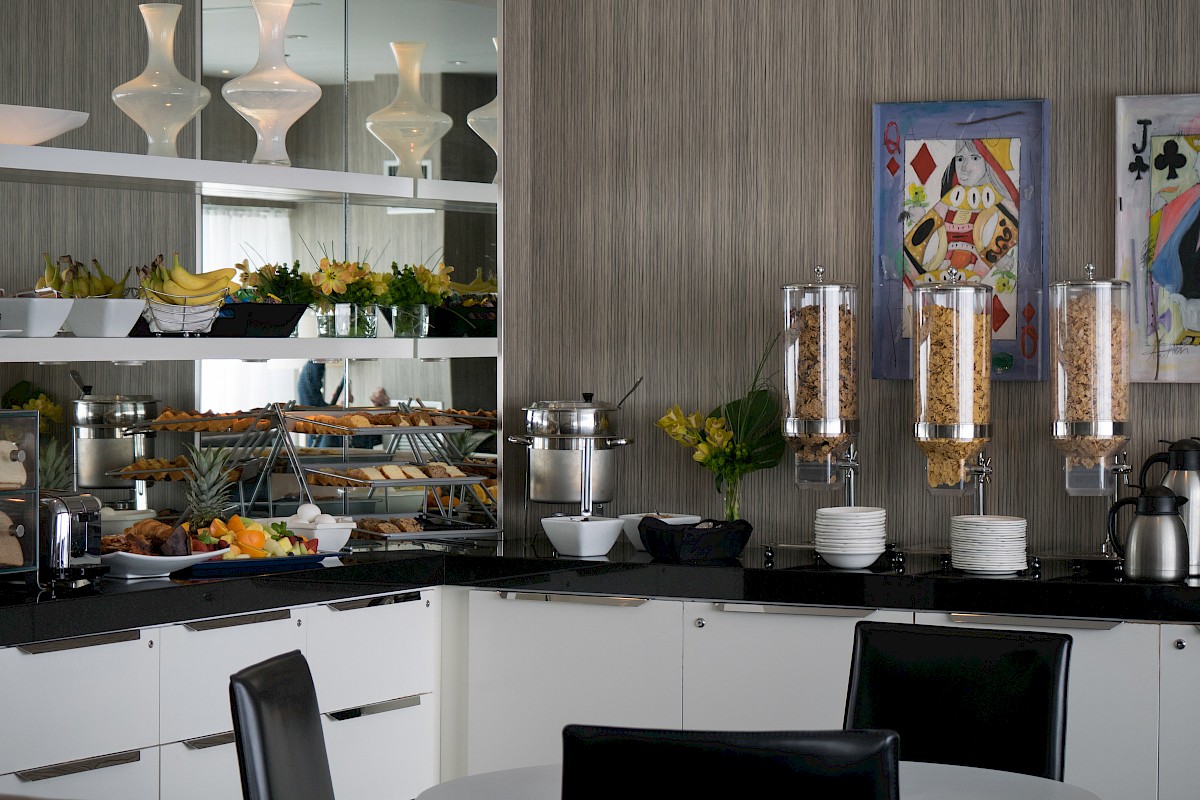 A modern dining area with a breakfast buffet, cereal dispensers, fruits, pastries, and decorative art on the wall completes the scene.