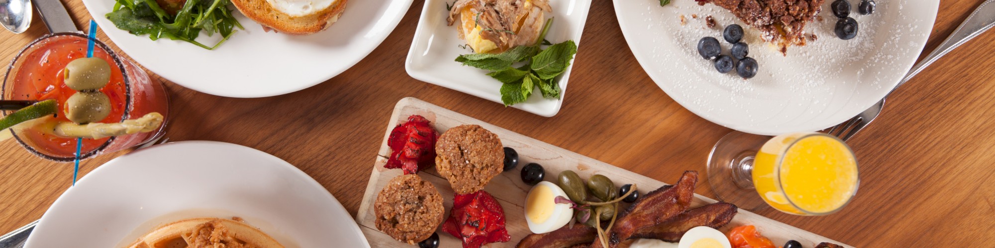 An assortment of breakfast dishes: waffles with chicken, fried egg on toast, pastries, salad, and drinks, displayed on a wooden table.