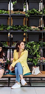 Two people sit on a bench with luggage near a vertical garden wall, in a bright interior space. One is on a phone.