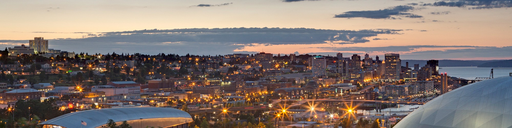 A cityscape during sunset with a skyline, buildings, and a brightly lit sports dome are visible under a partly cloudy sky.